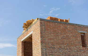 A brick structure stands incomplete with wooden beams positioned on its roof under a bright blue...