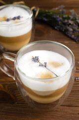Two cups of hot lavender latte on a white wooden table with flowers in the background. Concept of high-quality coffee drinks. Vertical orientation. Selective focus