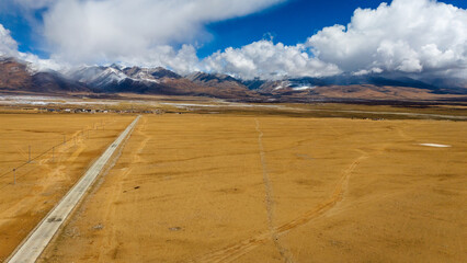 An empty road stretches across a vast and barren high-altitude plateau, leading toward snow-capped...