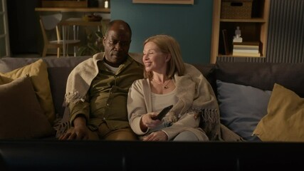 Caucasian mature woman changing channels holding remote control while watching TV with Black senior husband at home - Powered by Adobe
