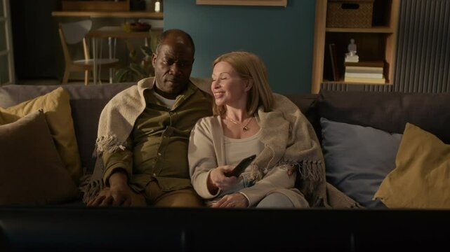 Caucasian mature woman changing channels holding remote control while watching TV with Black senior husband at home