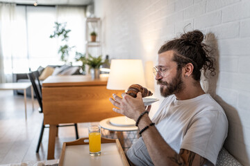 Young man enjoying breakfast in bed with croissant and orange juice