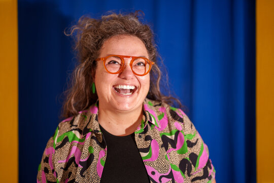 Cheerful female designer with curly hair, orange glasses, laughing while glancing sideways against vibrant blue and yellow backdrop
