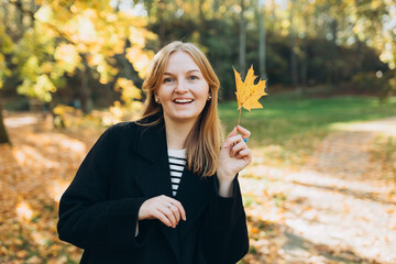 Portrait of pretty young 30s woman in evening autumn sun, smiling. Autumn landscape, calmness, tranquility concept. Pretty female holding leaf on nature background. High quality photo.
