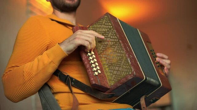 Close-up of man in mustard sweater playing accordion indoors, focusing on hand and buttons, warm lighting, cozy atmosphere, perfect for music and lifestyle stock footage.
