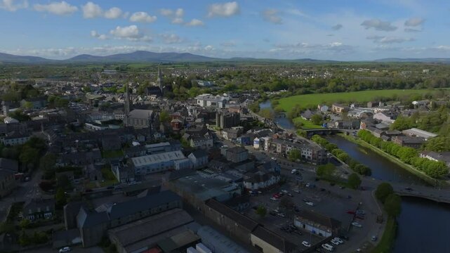 Enniscorthy, County Wexford, Ireland, April 2025. Drone high angle overview approaches above town covered by cloud shadow darkening the buildings and light breaking through onto church and castle.