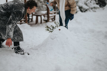 Two people enjoy a winter activity, making a small snow sculpture while dressed warmly, surrounded by snow-covered surroundings.