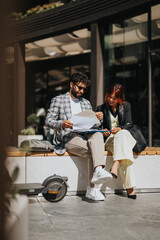 Two business professionals discuss documents while seated outdoors, exuding teamwork and collaboration on a sunny day.