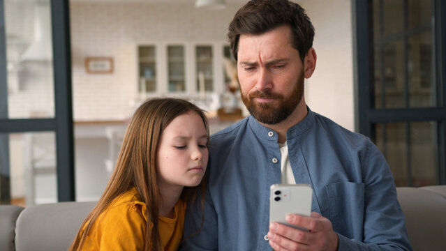 Disappointed Caucasian family guy man parent father holding mobile phone bad result girl kid problem negative sms at home upset child little daughter looking screen male dad failure online tech error