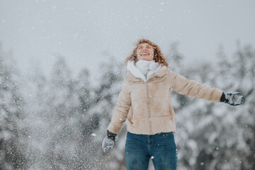 A cheerful woman dressed in a winter jacket and gloves enjoys the snowfall in a picturesque forest...