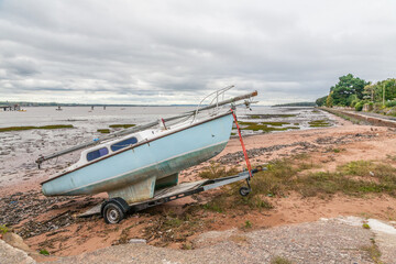 A weathered blue sailboat sits on the muddy shoreline. Signs of neglect and coastal erosion create...