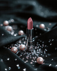Beautiful lipstick in shimmering pink shade is elegantly placed on black satin fabric, surrounded by pearls and tiny crystals, creating luxurious and glamorous atmosphere
