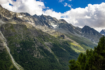 Naklejka premium Scenic view of rugged alpine mountains in Switzerland with rocky peaks, green slopes, and scattered trees under a blue sky with white clouds.