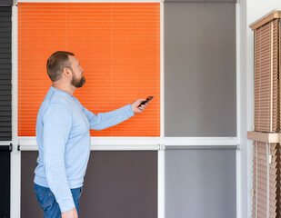 A man is in the showroom of a sun protection system. Demonstrates the operation of automatic blinds using a remote control, standing in front of various window coverings including pleated blinds.