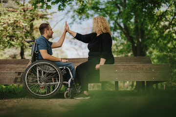 Man in a wheelchair high-fiving a woman on a bench in a park, evoking a sense of teamwork,...