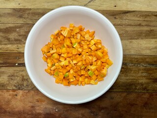 photo of carrot slices cut into small squares then placed in a white plastic bowl with a dark brown wooden table background, carrot slices ready to be processed into delicious dishes