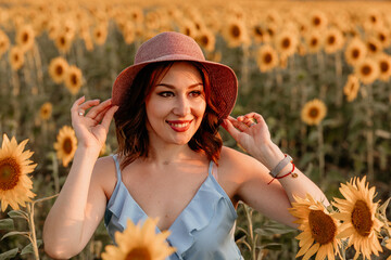 Beautiful happy young girl in blue dress and hat in sunflower field at sunset