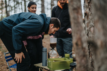 Three friends enjoy a camping trip in the forest, engaging in an outdoor activity of making coffee using a siphon coffee maker on a portable table surrounded by trees.