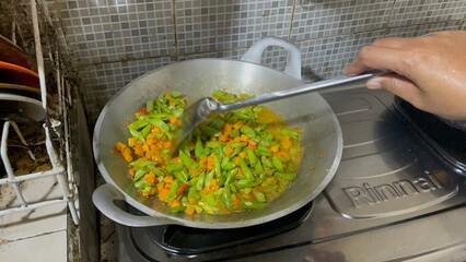 housewife is cooking stir-fried tempeh, green beans and carrots using a frying pan and spatula on the stove, a dish suitable for serving as breakfast