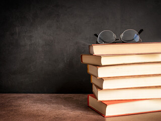 Stack or pile of hardcover books with eyeglasses on top on dark brown table against blackboard...