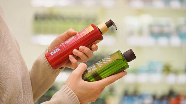Customer comparing haircare products in a beauty store. Woman reading ingredient labels on bottles of shampoo, conditioner or balm. Concept of conscious choice and personal care for hair and scalp.