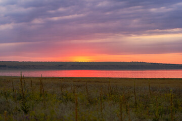Obraz premium Colorful summer sunset over river with cloudy sky and meadow in foreground, dramatic warm light reflecting in the water near coastal hills