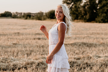 Beautiful happy young blonde girl in a short white dress in a field at sunset