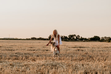 Beautiful happy young blonde girl with labrador in field at sunset