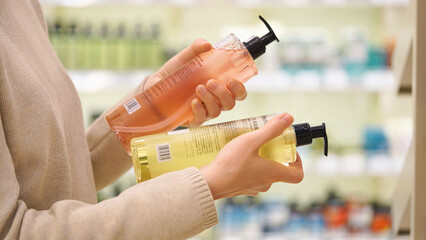 Customer compares haircare, personal care, skincare products in a cosmetics aisle. Woman reads labels and examines the ingredients. Closeup female hands holds two different bottles. Conscious choice.