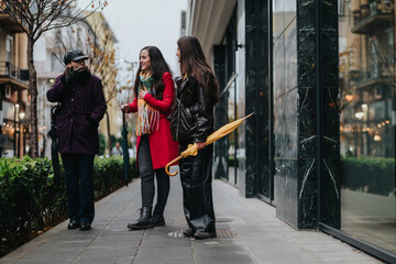 Three women dressed in winter attire engage in a friendly conversation while walking through a city street, showcasing urban life and casual gatherings on a slightly overcast day.