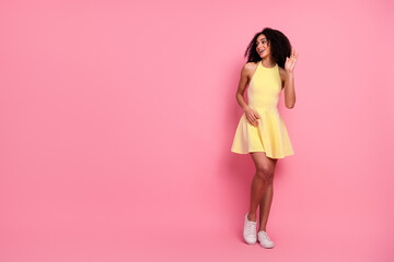 Young woman in a vibrant yellow dress and sneakers with stylish curly hair posing against a pink...