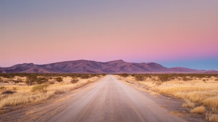 Desert road leading to mountains under a pastel sky at sunset with dry grass on either side of the road