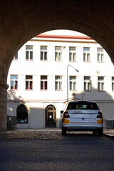 White medical transport car with red and yellow markings parked under stone archway near building. Historic brick arch frames vehicle, street signs nearby, cobblestone pavement, warm sunlight highligh