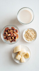 A bowl of mixed dried fruits, a plate of sliced bananas, a bowl of oats, and a glass of milk arranged on a white surface.