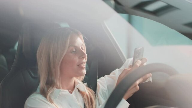 Content blonde woman holding phone in one hand while touching smartwatch with other arm resting on wheel. Checking time or syncing device. Managing tasks efficiently during relaxed pause in car.