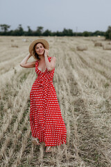 Beautiful young girl in a red polka dot dress and hat in a field of haystacks