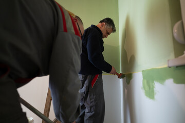 Two men are engaged in painting a room's walls with green paint, paying close attention to detail....