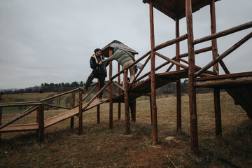 Two individuals interact on a wooden structure in a scenic rural landscape during overcast weather,...