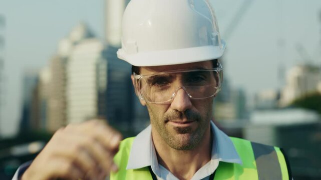 Portrait of man engineer in protective eyewear putting on safety helmet while looking at camera. Male builder in reflective vest adjusts hardhat preparing for construction tasks