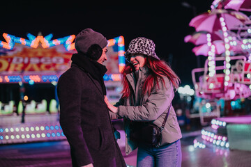 A cheerful couple laughing together while visiting an amusement park at night, filled with colorful...