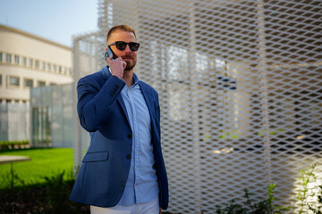 Businessman wearing sunglasses is making a phone call after work, standing in front of a modern architectural structure in an urban setting, enjoying the pleasant weather