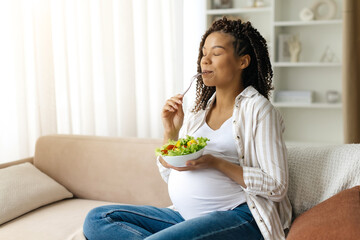 Black pregnant woman eating fresh vegetable salad at home, young african american female enjoying...