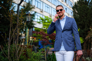 Smiling businessman wearing sunglasses is using smartphone while leaving office building after work, carrying briefcase and walking near green trees