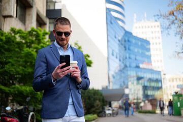 Young businessman wearing sunglasses, enjoying coffee while using a smartphone outdoors near modern office buildings, embracing a stylish and confident lifestyle after a busy workday