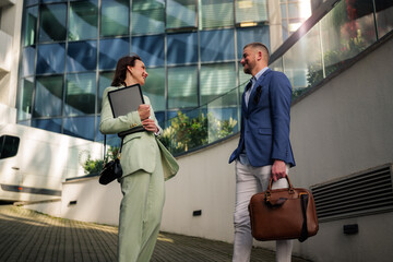 Two business professionals are having a conversation outside their modern office building after work, possibly discussing project updates or sharing insights, while holding a tablet and a briefcase