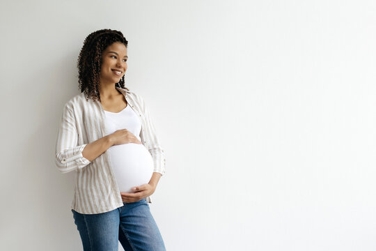 Black pregnant woman gently touching belly, looking away and smiling. Peaceful and uplifting maternity portrait on clean white background with natural light, copy space - Powered by Adobe