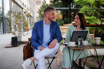 Two business colleagues are sitting at an outdoor cafe, enjoying a coffee break after work, discussing projects, and relaxing together in a friendly atmosphere