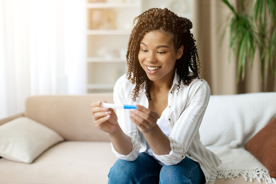 Happy black woman smiling while holding pregnancy test. Represents joy, surprise, and excitement after discovering she expecting in bright home environment, copy space - Powered by Adobe