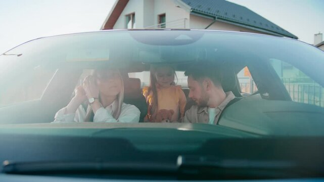 Blonde woman checking mirror and fixing hair before departure. Bearded man focused on phone setting route. Little girl in yellow holding plush toy and hugging father. Starting family drive from home.