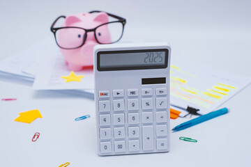 Finance and budget planning concept with a pink piggy bank wearing eyeglasses, a calculator displaying 2025. Documents with highlighted text, and colorful office supplies on a white desk.